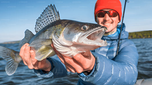 A person holding a freshly caught fish with a smile on their face, wearing a red cap and blue jacket, with the ocean in the background.