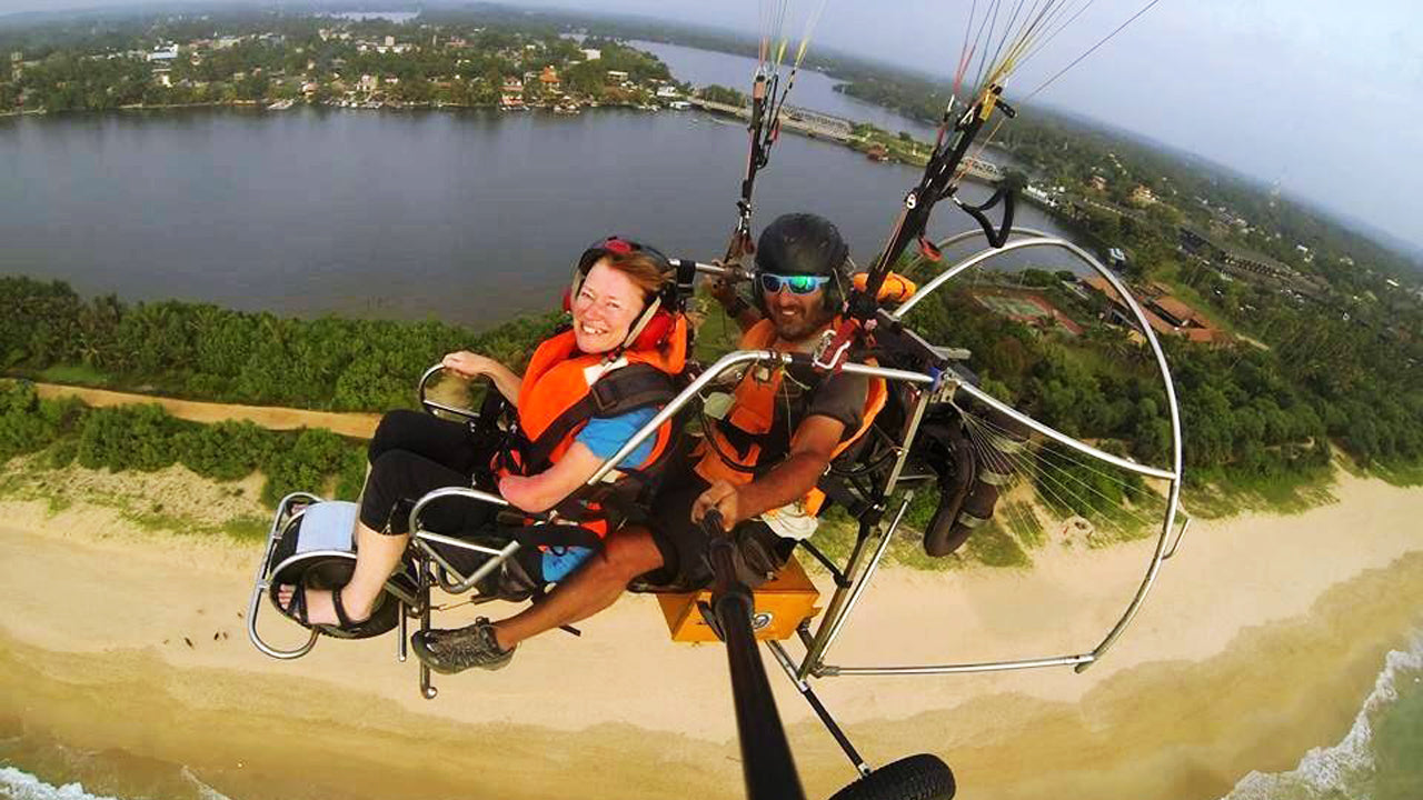 Two people in a hang glider over a beach with a lake and trees in the background