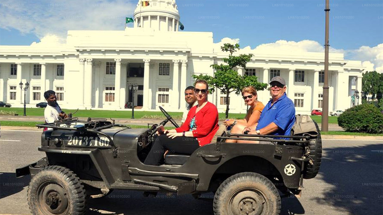 A group of tourists riding in a green and black vintage war jeep with the roof top down, passing by a white building that resembles a governmental or historical building.